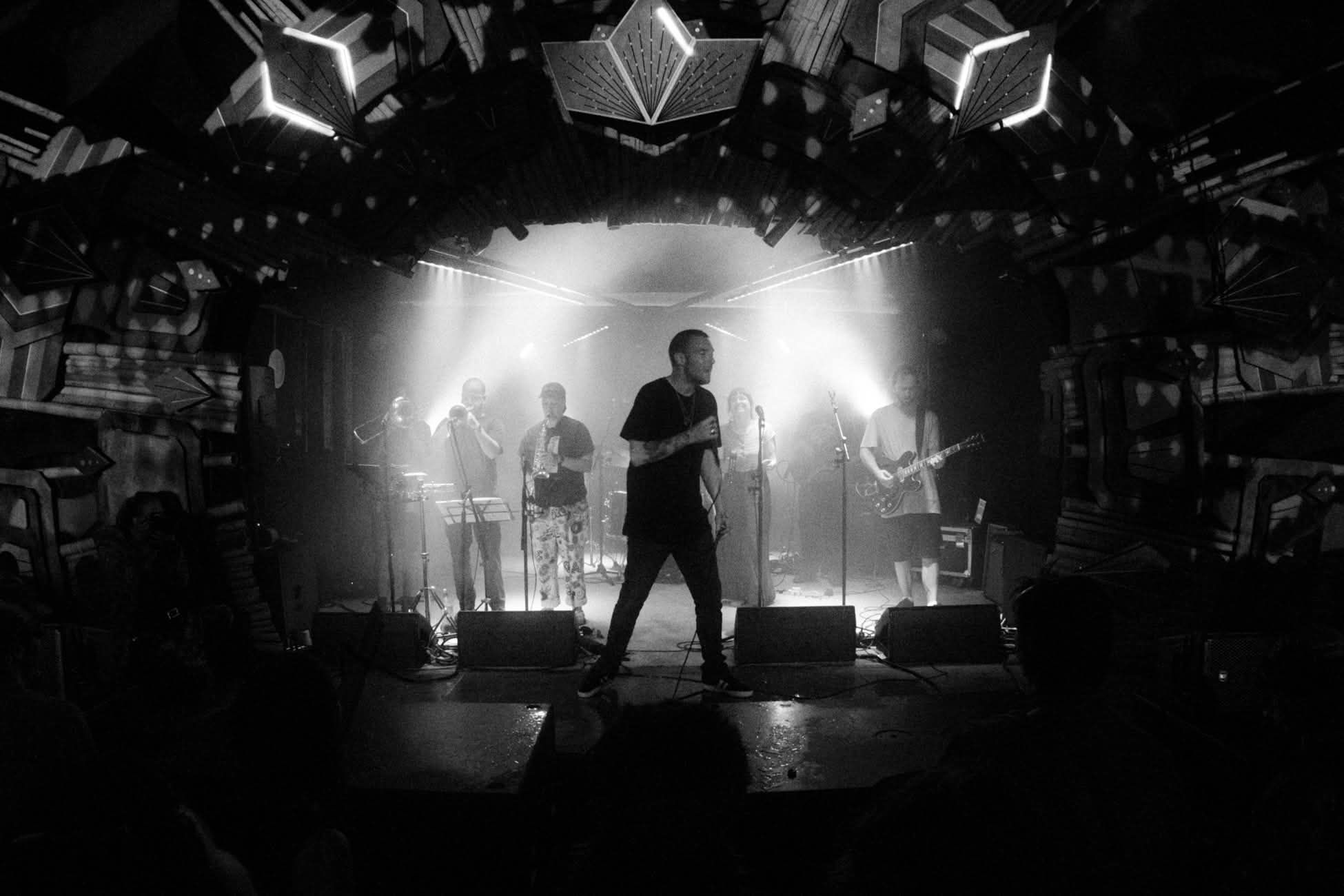 A black and white image, the singer of the band is silhouetted starkly at front of stage and the band are in a pool of bright behind them