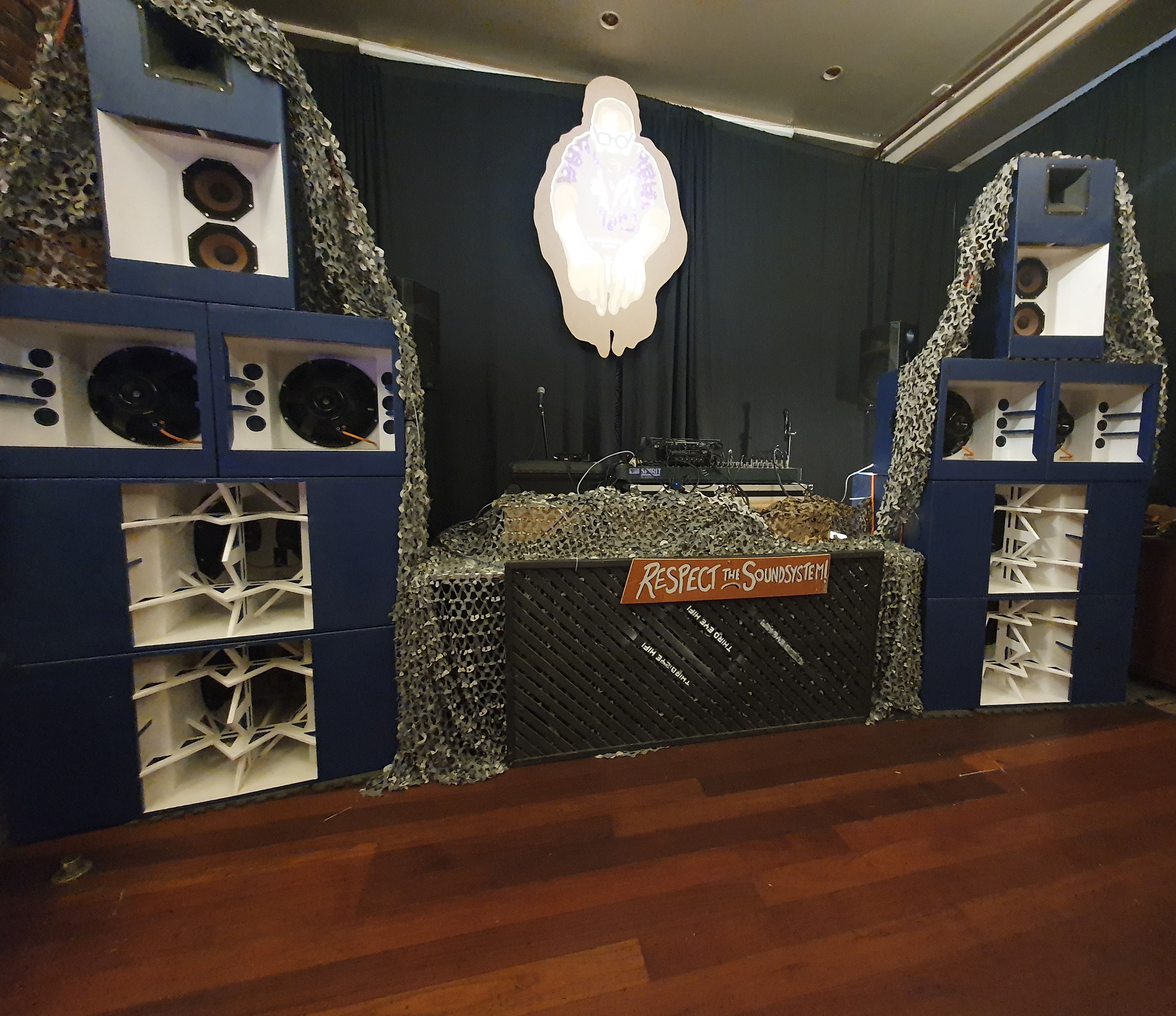 Two speaker stacks in an indoor venue, with a &#39;respect the soundsystem&#39; sign visible hanging between them in front of the DJ booth