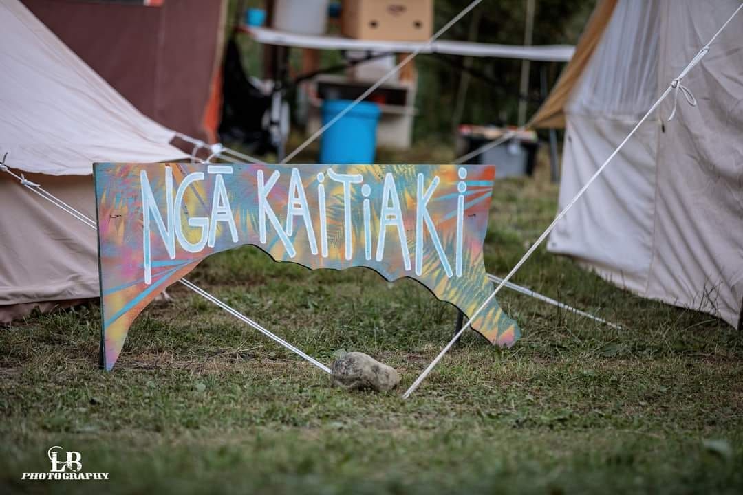 The Ngā Kaitiaki sign on a funky piece of wood in a paddock, with tents and tables visible behind