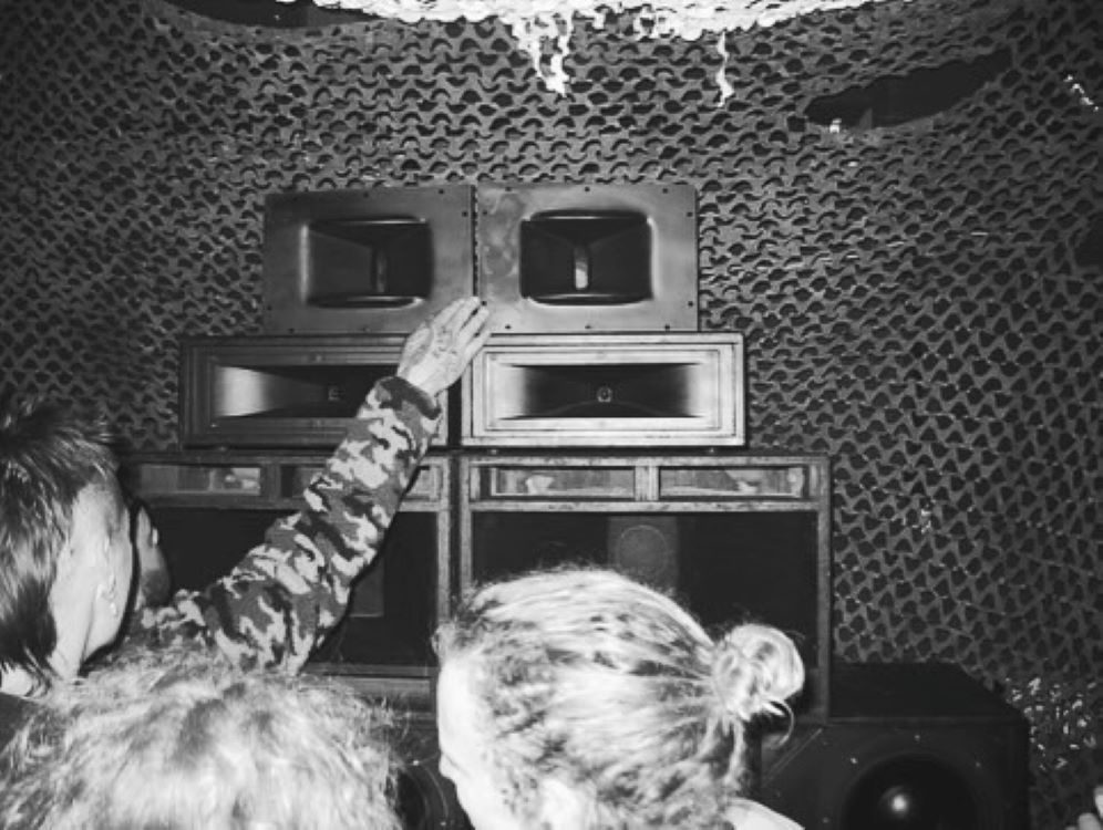 A black-and-white image of a couple of people dancing, in front of a soundsystem, with a cargo net as backdrop