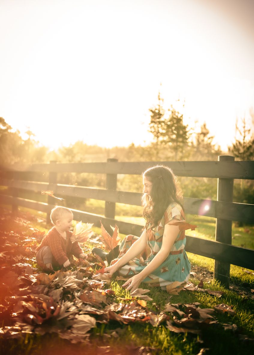 A wholesome image of a toddler and adult playing in autumn leaves, next to a stout wooden fence, with trees in the background