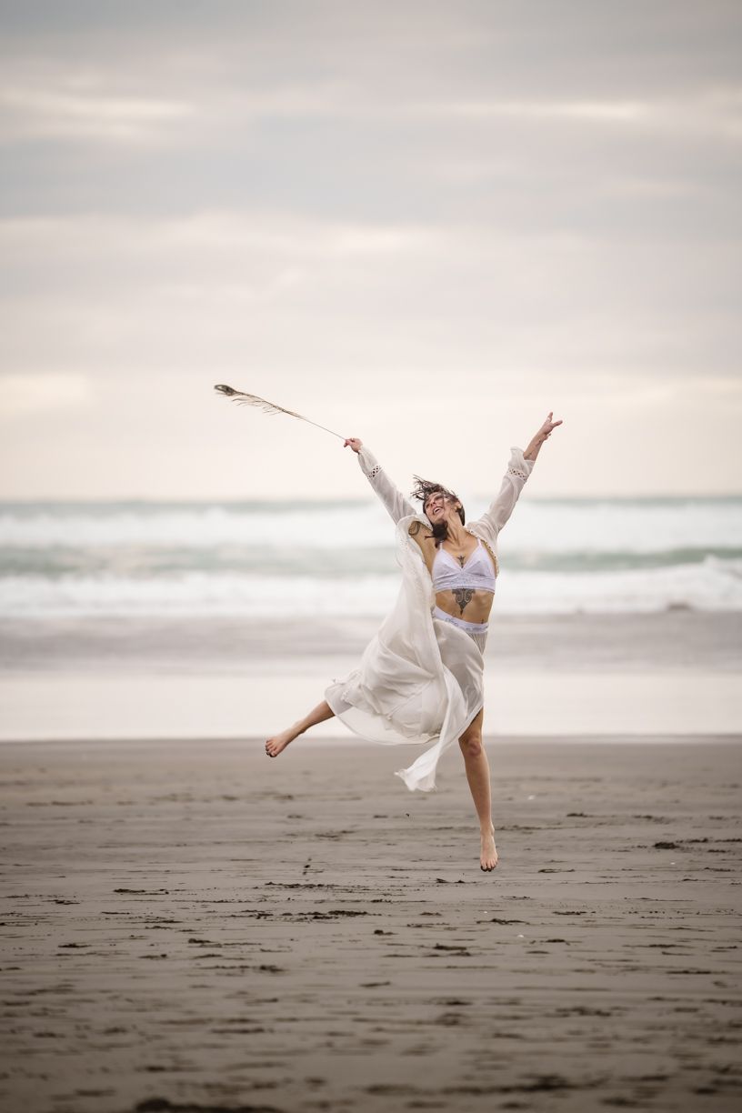 A person mid-leap on a wide beach, the white of their flowing outfit matches the grey of the sky and sand and white of the rolling waves behind them