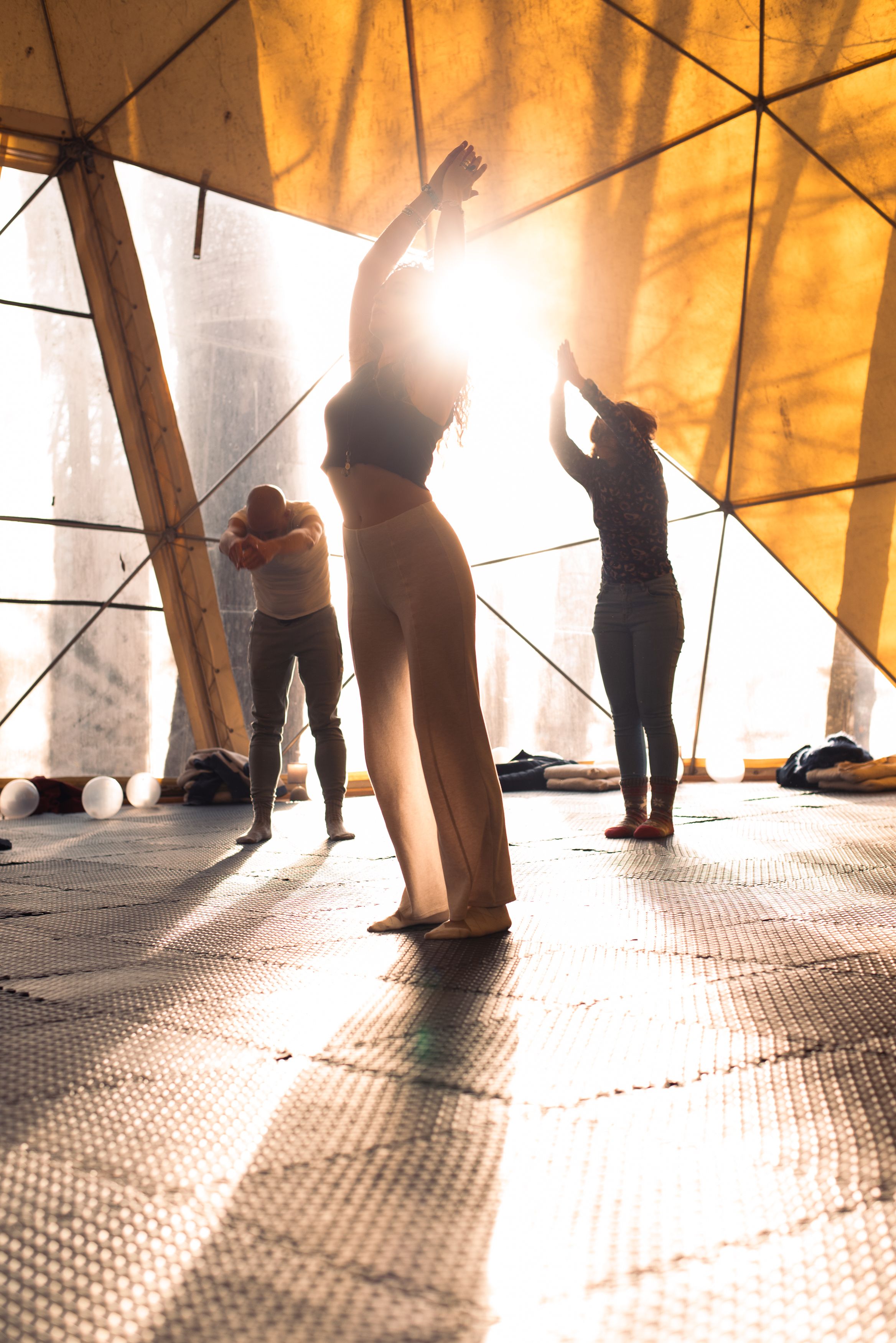 Three people stretching their arms upwards in a covered outdoor area, the sun silhouetting them from behind