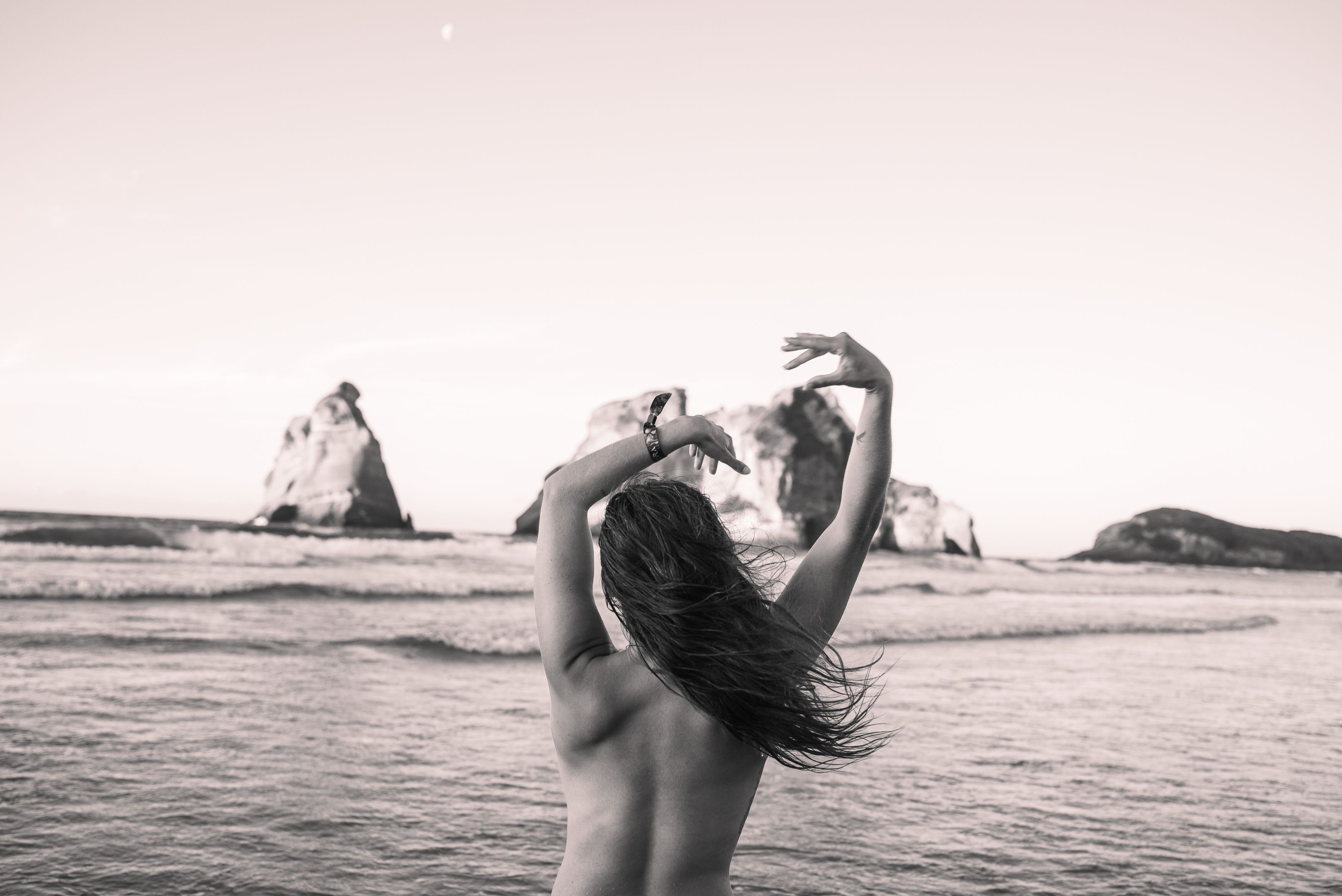 A black-and-white image, of a topless femme person&#39;s torso from behind, their hair is loose and arms raised. The ocean and jagged rock features are in the background. 