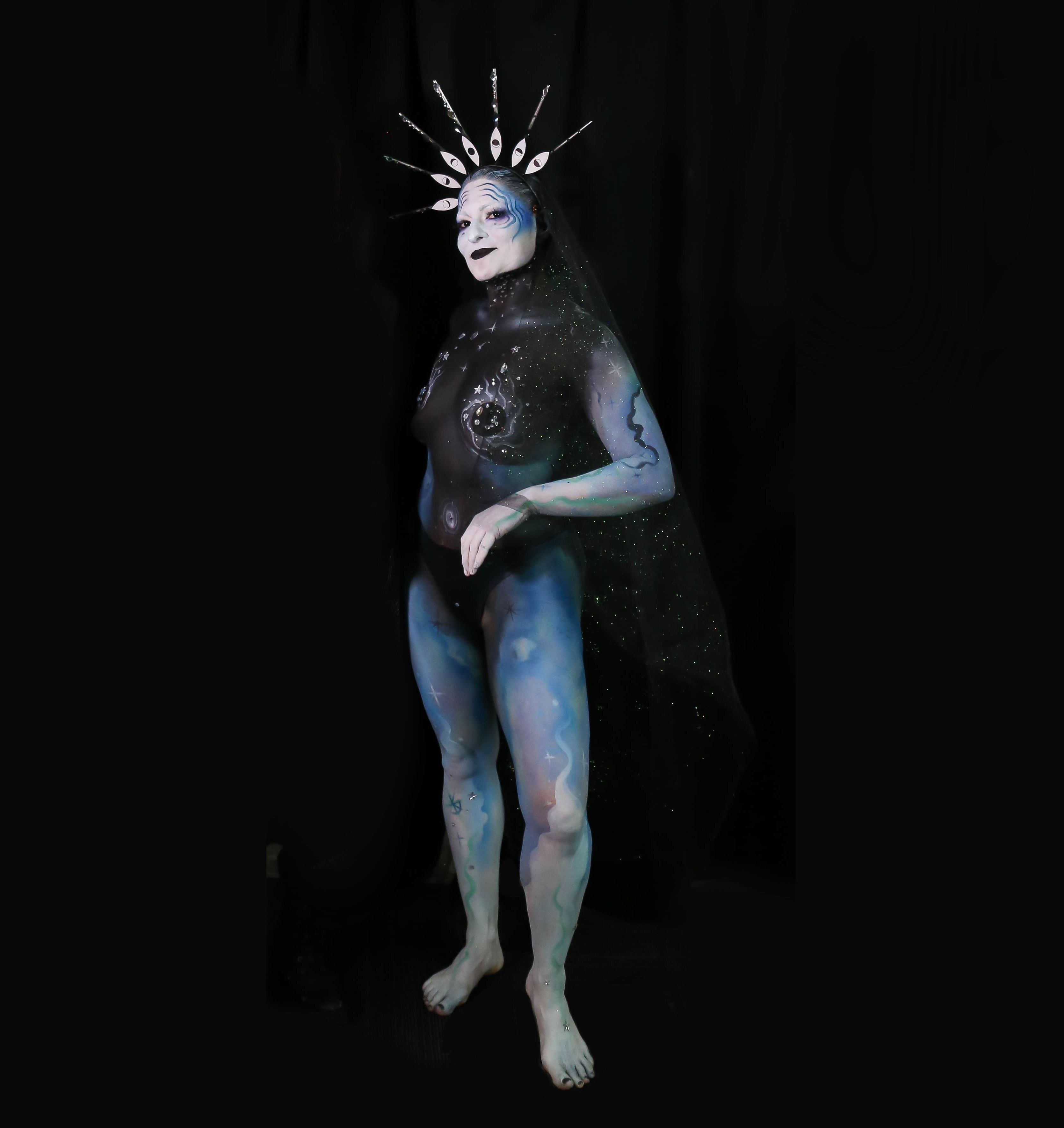 A person in full body paint, with wavy blue over their limbs and an incredible head-piece of eyes
