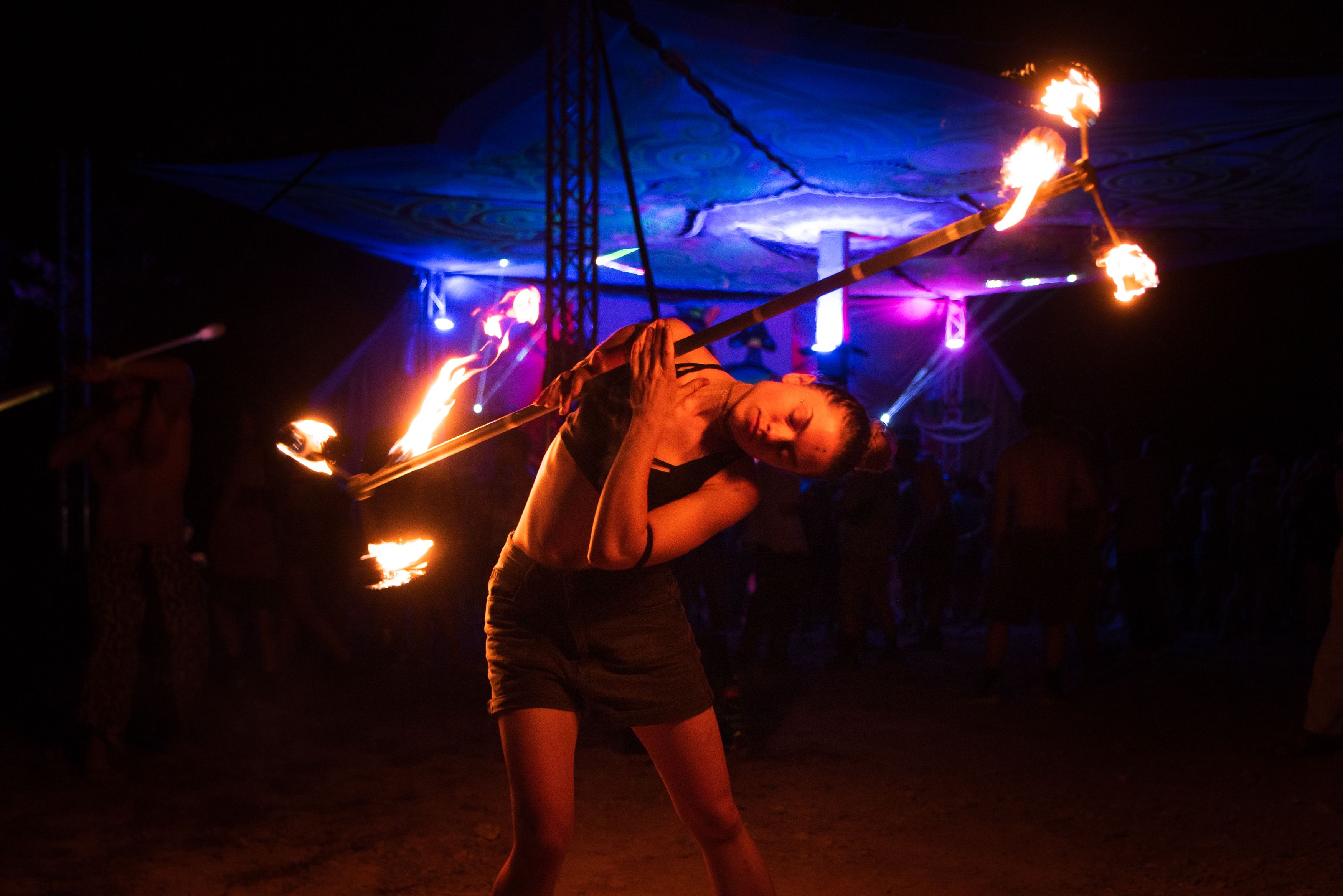 A firespinner leaning sideways to roll a dragon staff across their torso, with blue lighting against a canopy behind them