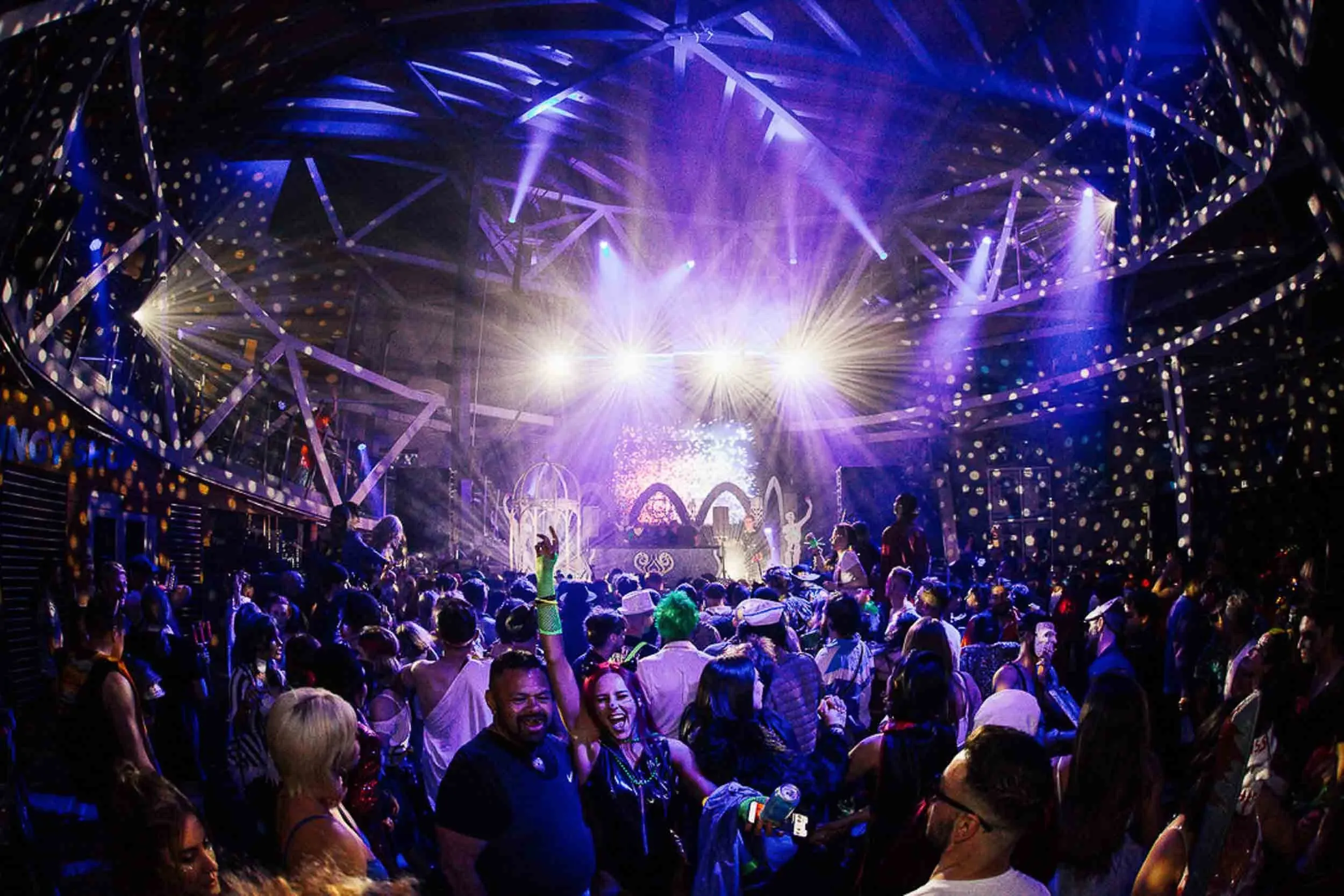 A fish-eye lens shot of an event, the stage is the focus with smiling crowdmembers dancing in the foreground, purple lights crisscross the image