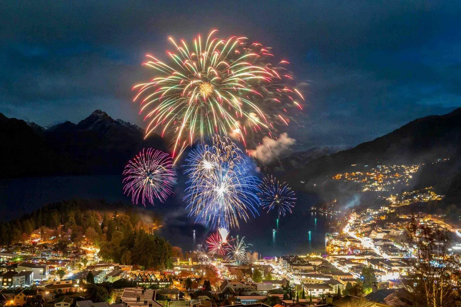 Fireworks against a dark sky, with mountains behind and warm town lights visible below them