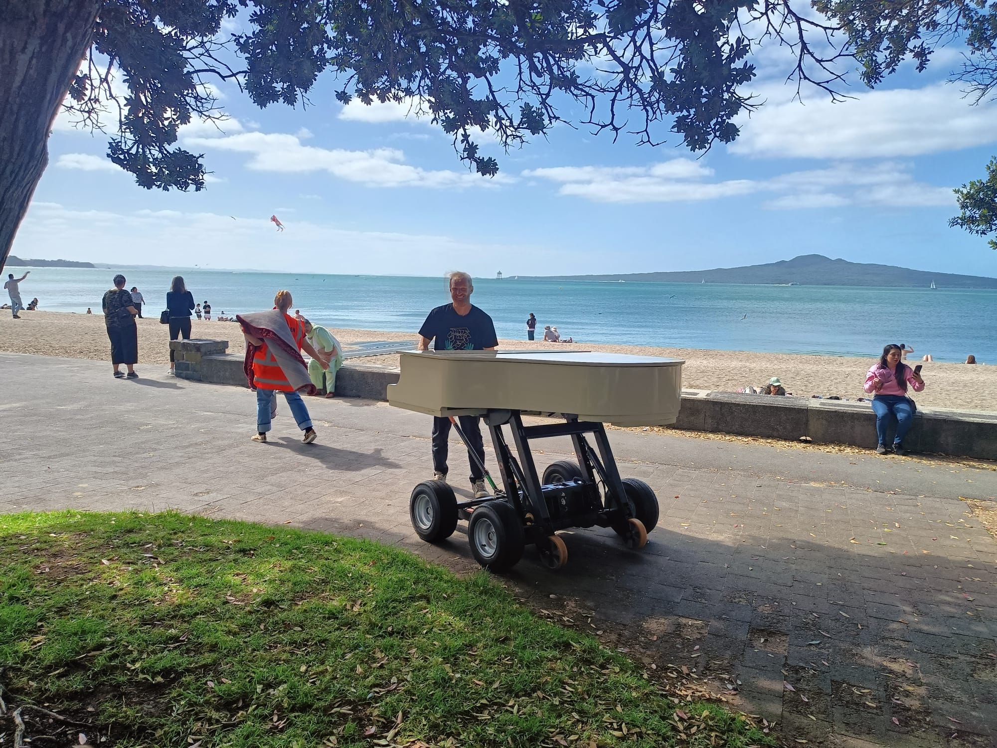 The piano on a trolley with chunky off-road wheels, on the path in front of a Tāmaki beach, Rangitoto in the background. A person is standing grinning behind the piano.