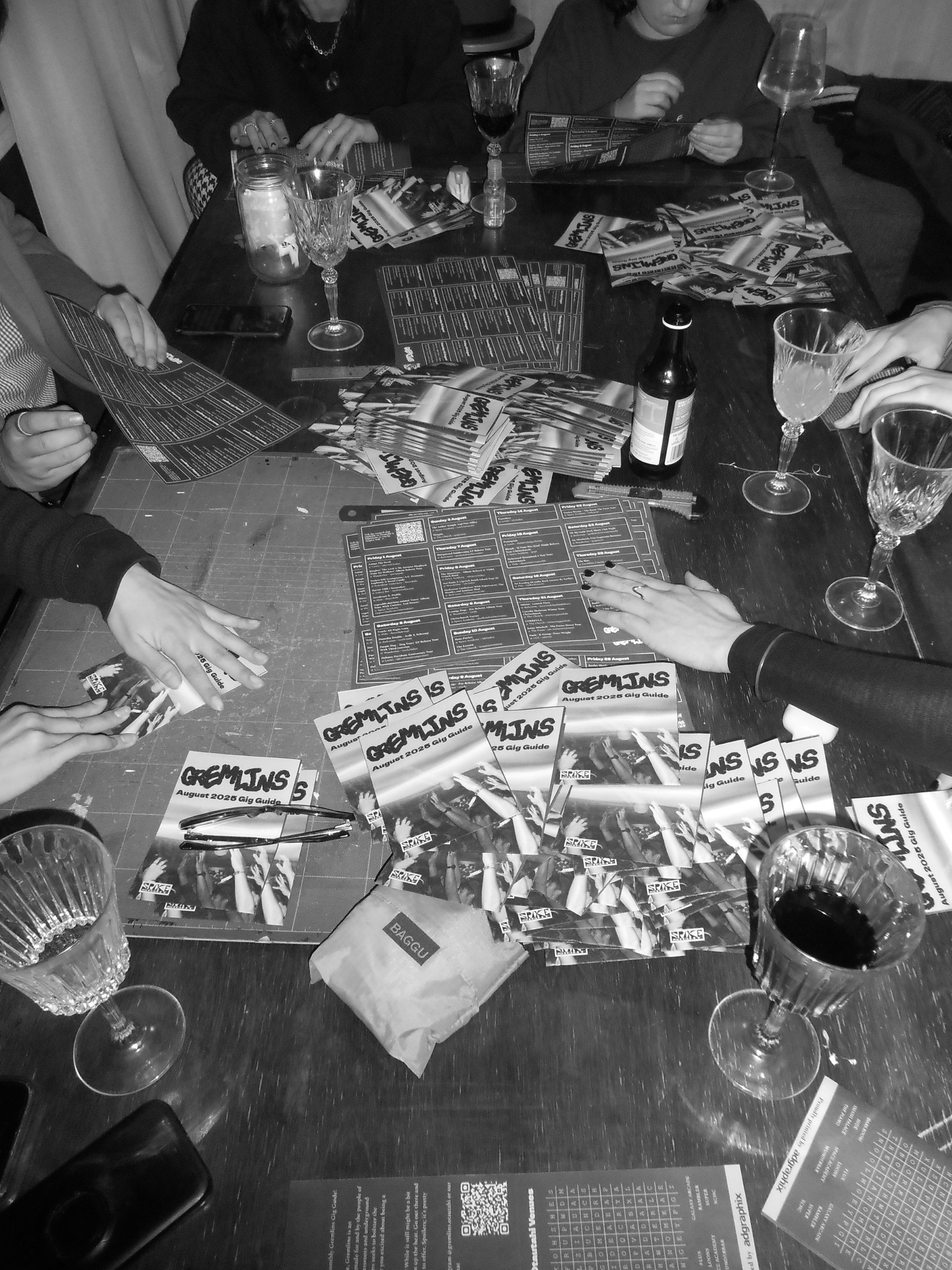 A black-and-white image of a group of people folding Gremlins zines or gig guides. The zines are piled on the table in between crystal glasses, phones and pens.
