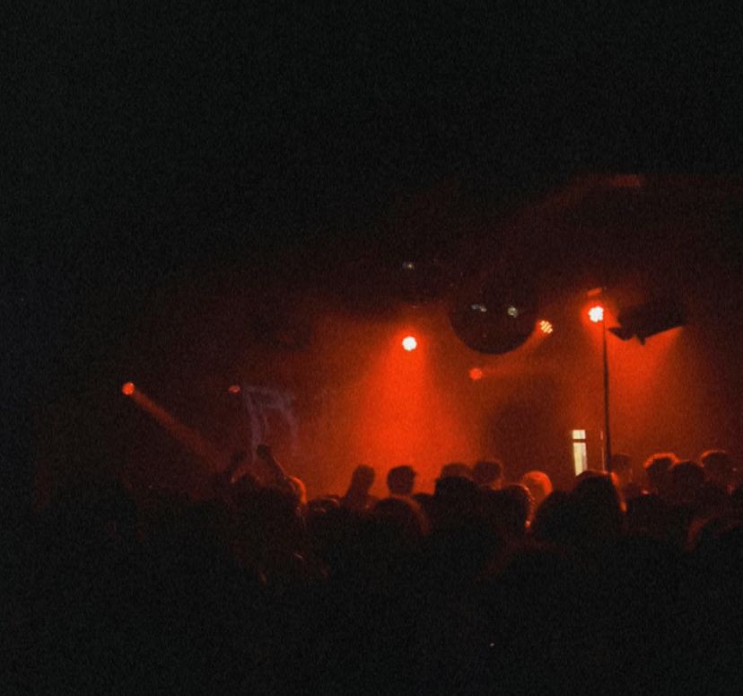 A dark room lit with warm red lighting, showing a crowd facing a stage