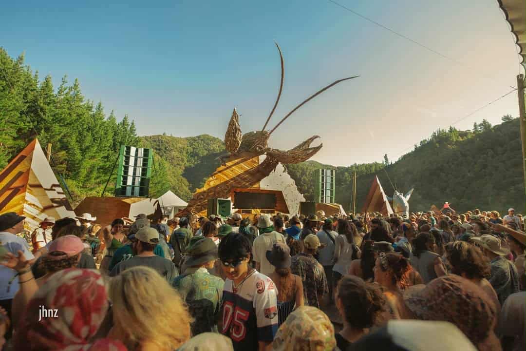 A long shot of a stagefront seen over the heads of a large crowd. The stage is overlooked by a huge driftwood kōura/freshwater crayfish