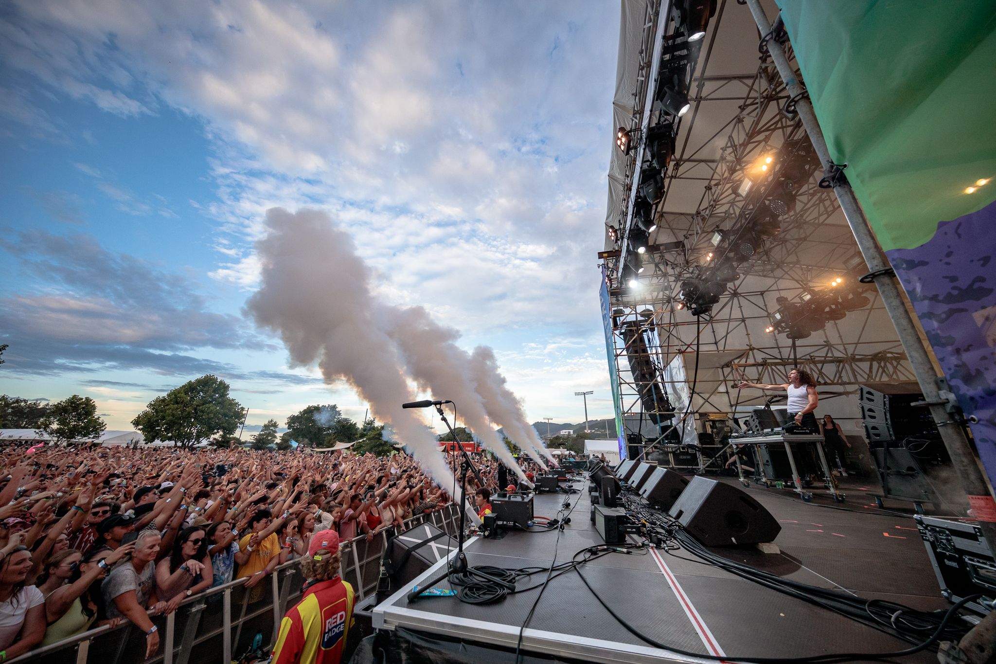 A side view of a large stage, with smoke cannons shooting up to a cloudy blue sky over a packed crowd