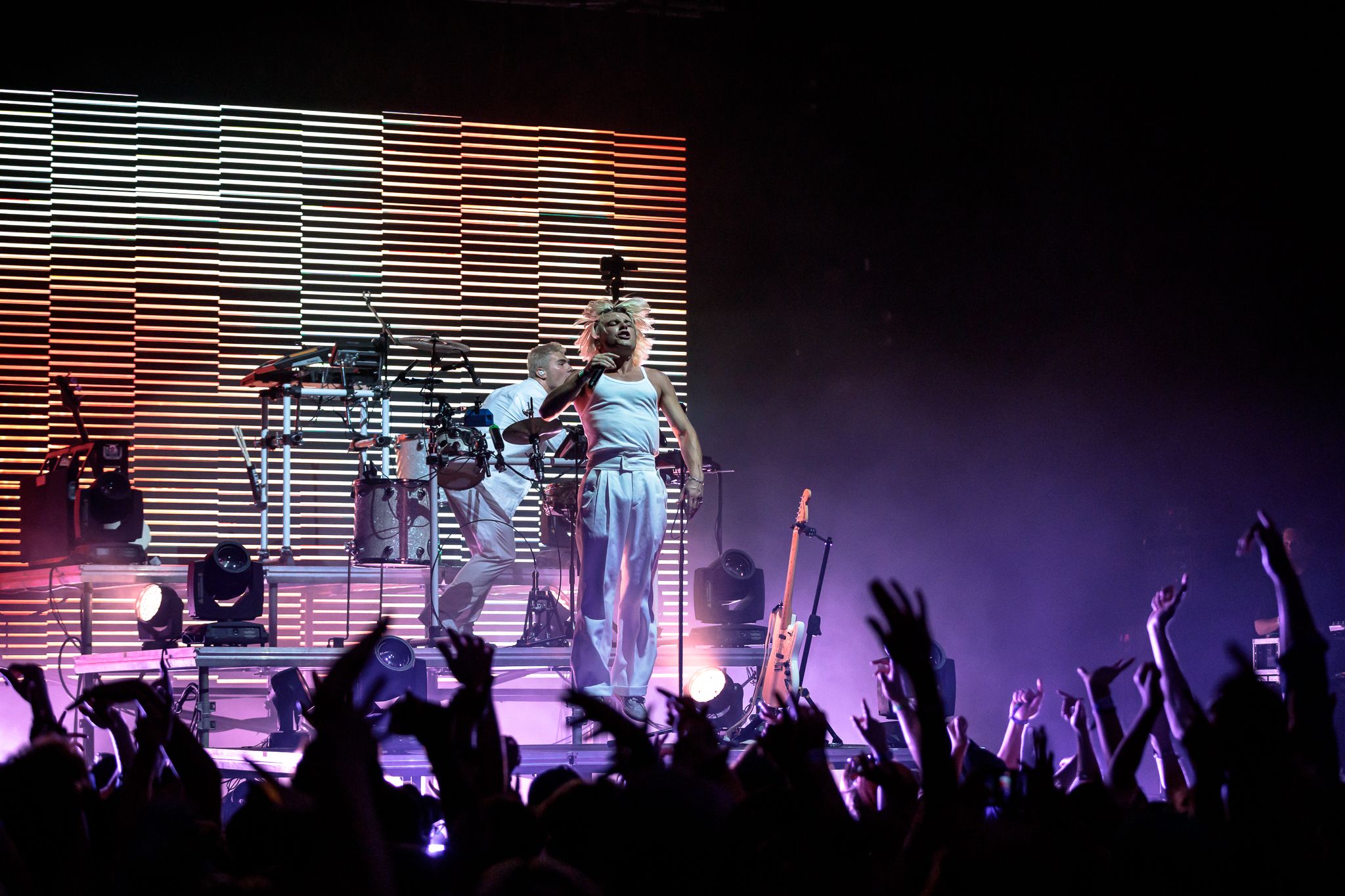 Two performers, one with a mic and one on drums, are facing the crowd. Silhouettes of hands raised are visible in the foreground.