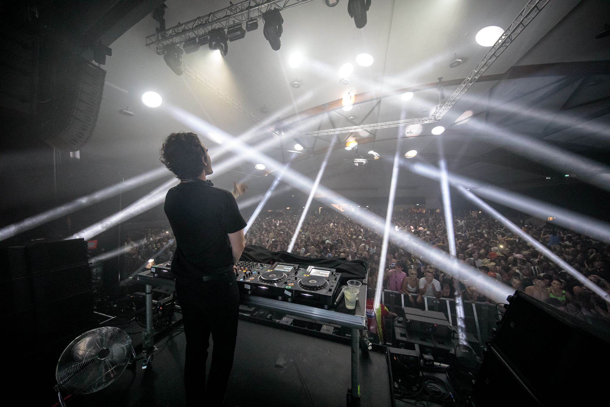 An image taken from the stage, over the shoulder of a performer wearing all black. Spotlights crisscross the huge crowd, adding to the black and white effect.