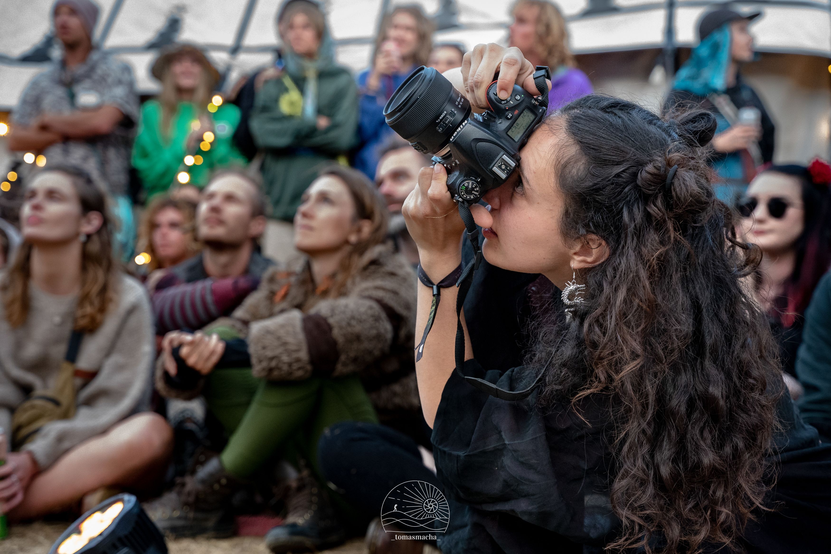 Marina in action, holding the camera to her face, shot from the side. A seated crowd are visible behind her