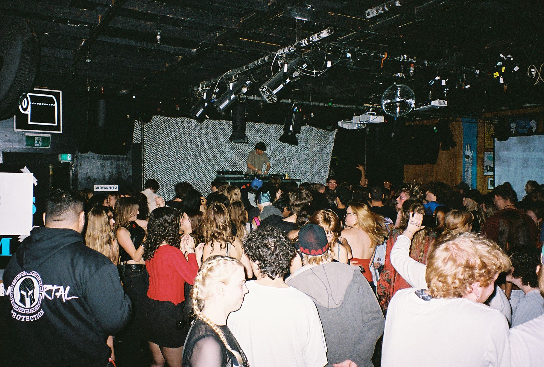 An indoor gig, photographed from the back across the full crowd. A DJ stands out against a staticky grey backdrop on the stage.