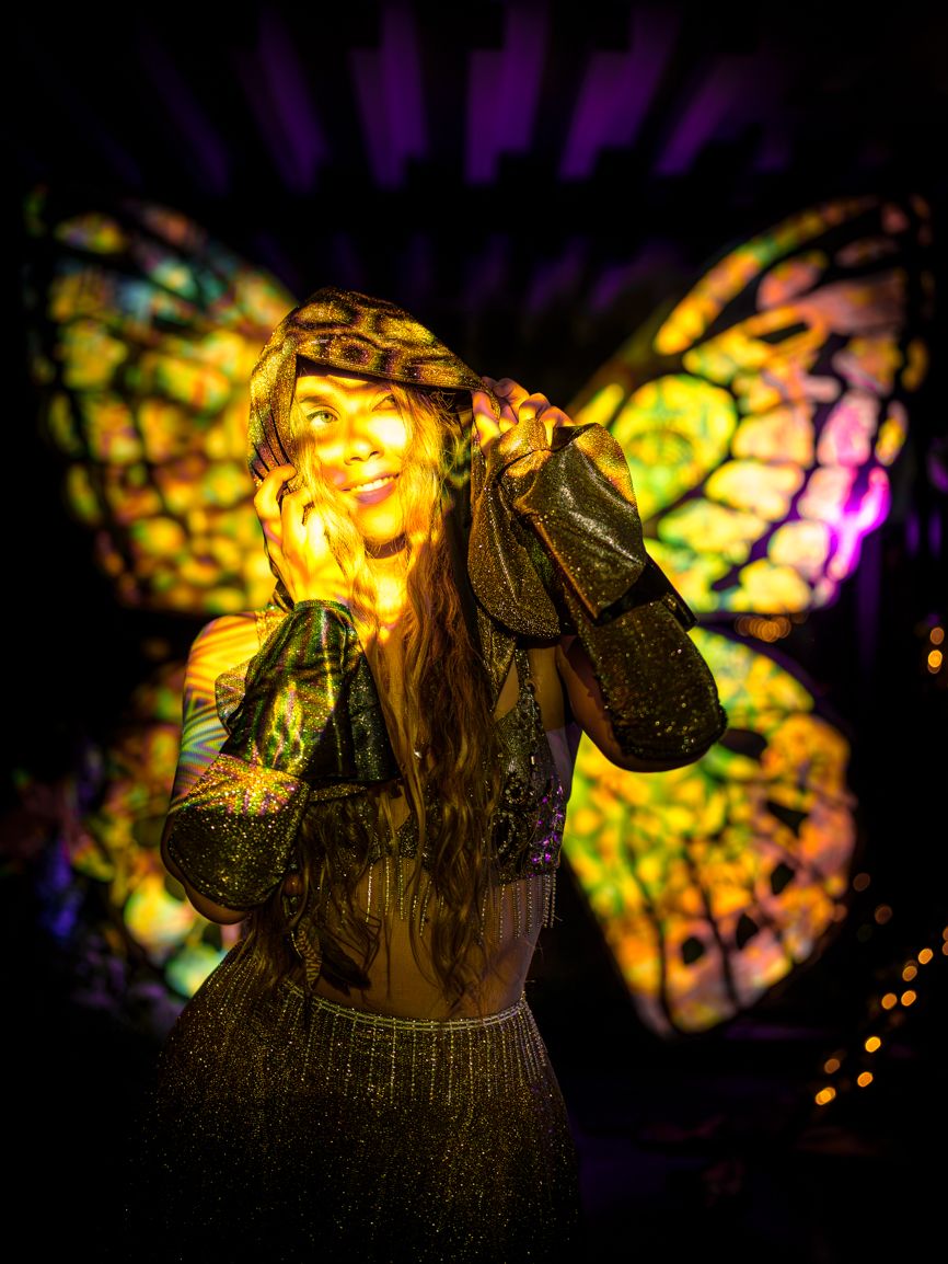 A person smiling past the camera, hands up to her hood, lit by colourful lights that light up a butterfly backdrop