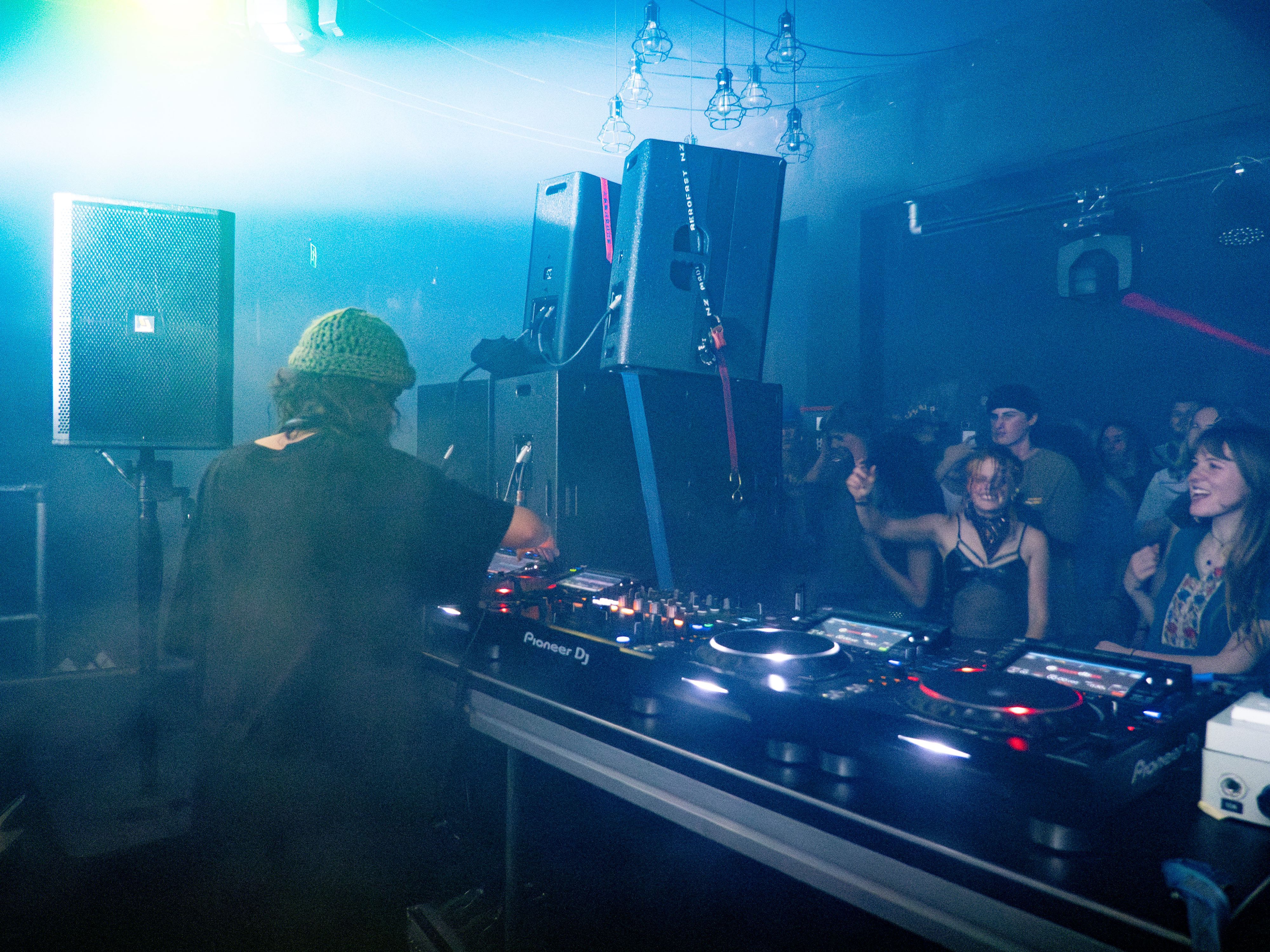A side-on shot of the DJ behind the booth, with the stacks visible in front of them, and the front row of the crowd throwing down
