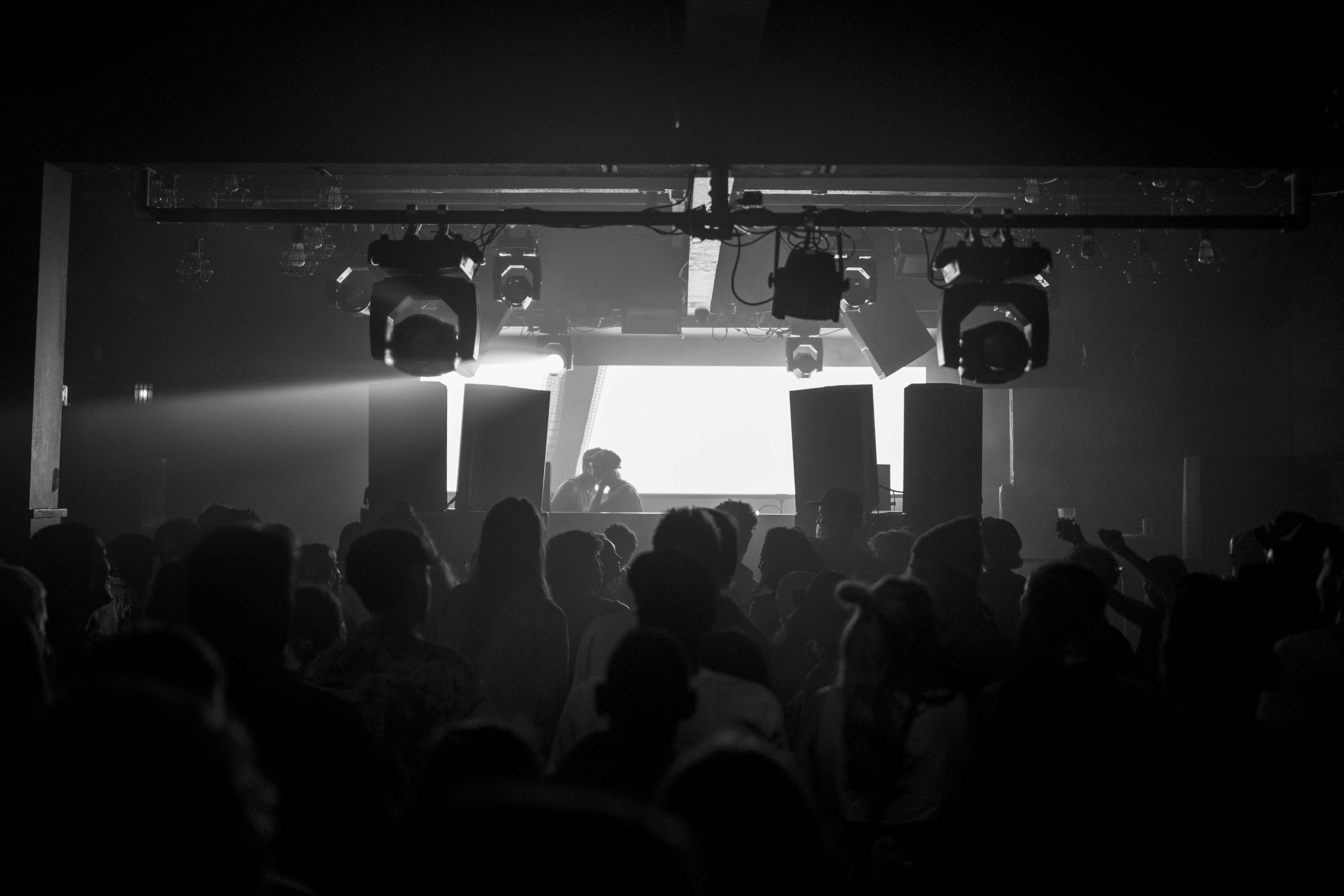 A black and white image, two sets of two speakers are aligned either side of the DJ, and the room is backlit by a large screen. A silhouette of a crowd is just visible in the foreground.