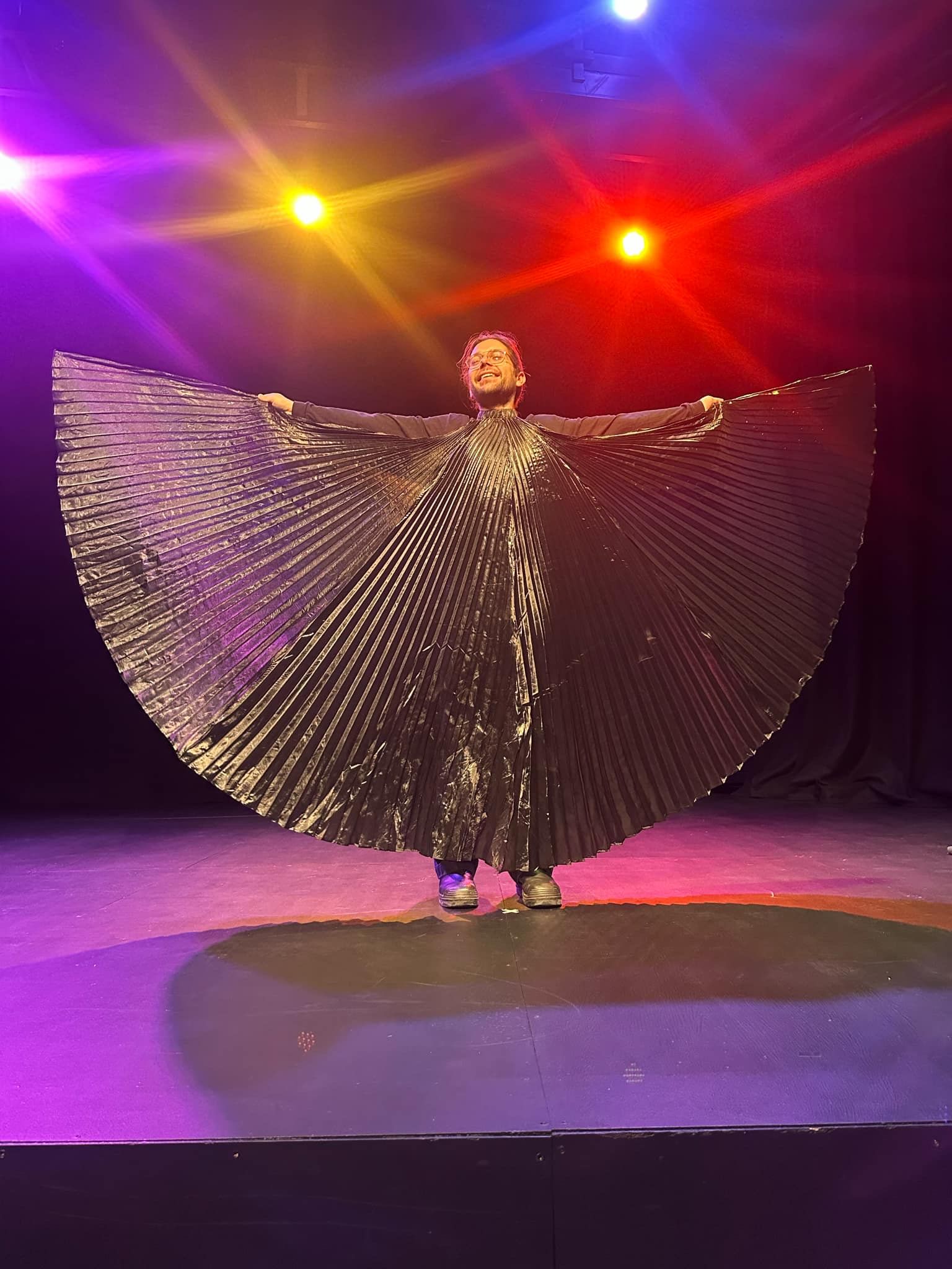Someone in a cape/wing suit, holding the cape wide and lit warmly from behind, on a stage