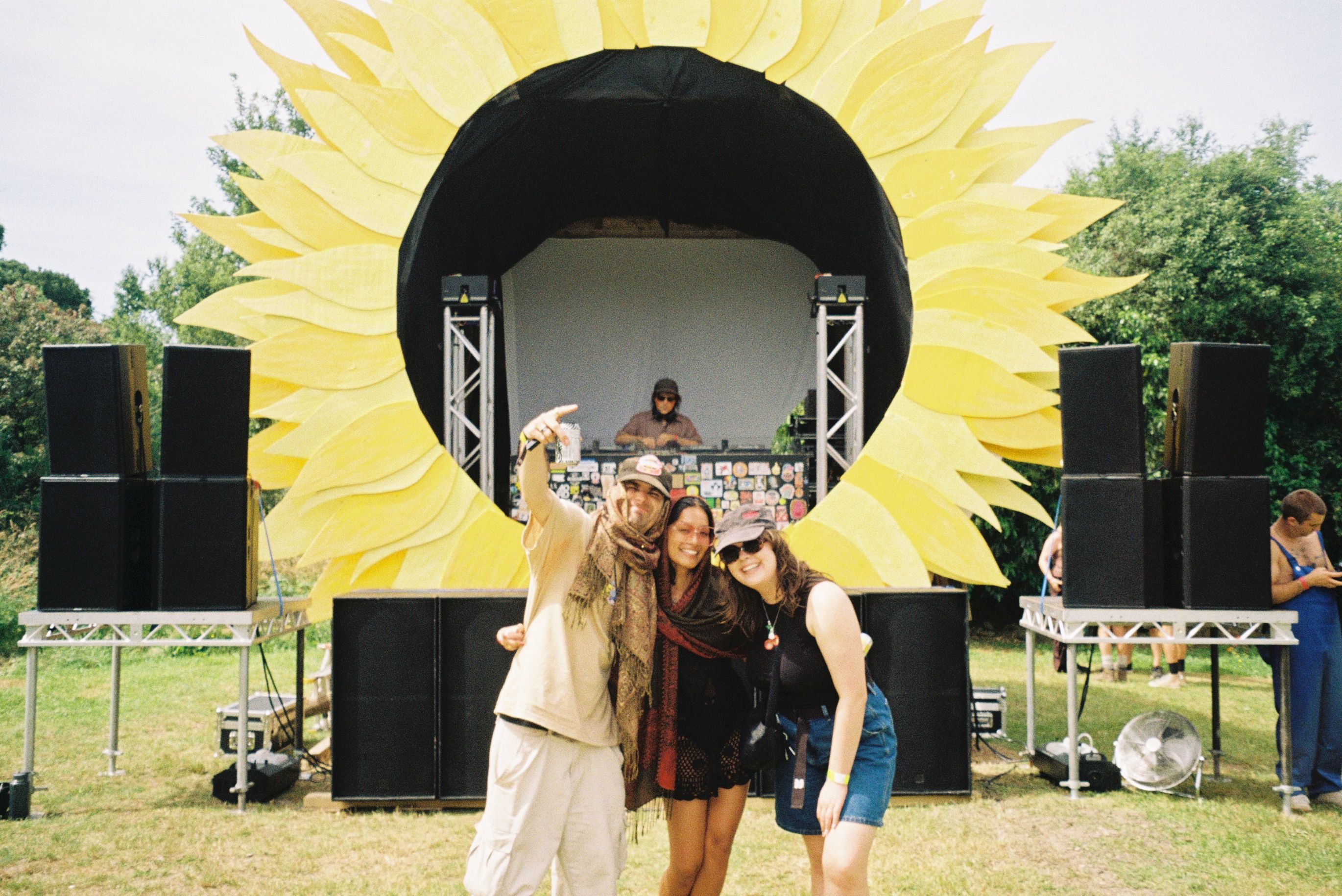 Three people pose in front of a sunflower-shaped stagefront, with speakers arranged along the stagefront. It's a bright day in a paddock.