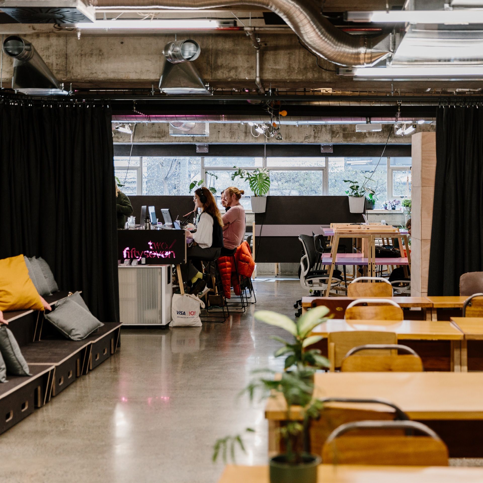 The co-working space, with empty tables visible in the foreground, and couple of people working at a tall table in the background. There are plants and colourful pillows dotted around the room.