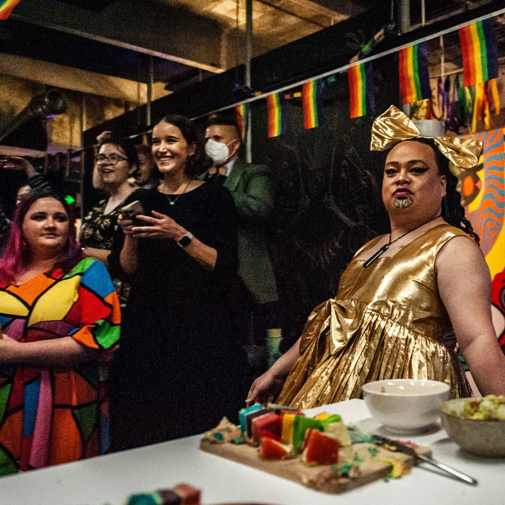 In the foreground is a wāhine with a moko kauae and a fantastic golden dress. Other people are standing just behind her. There are snacks on a table in front of them all.